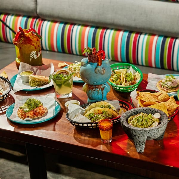 A table laden with various Mexican dishes, including tacos, tostadas, guacamole, and drinks.