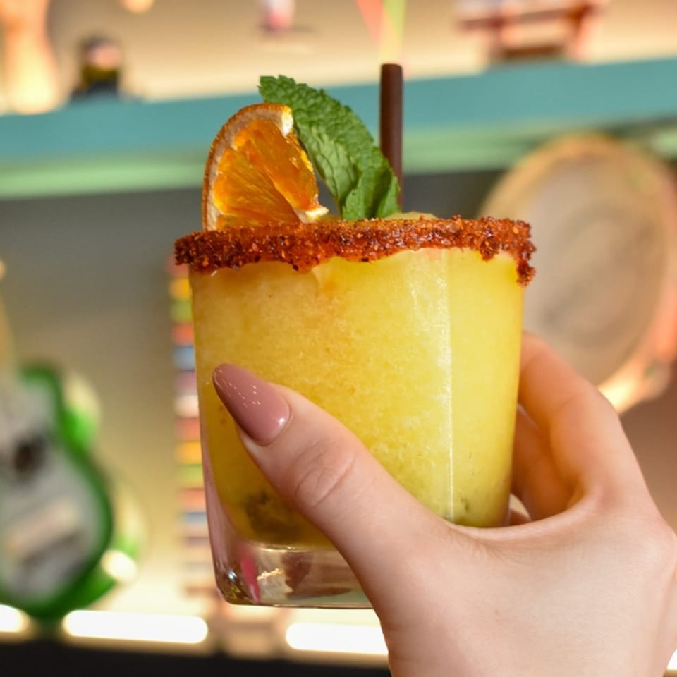 A hand holds a yellow frozen drink garnished with an orange slice, mint, and chili rim.