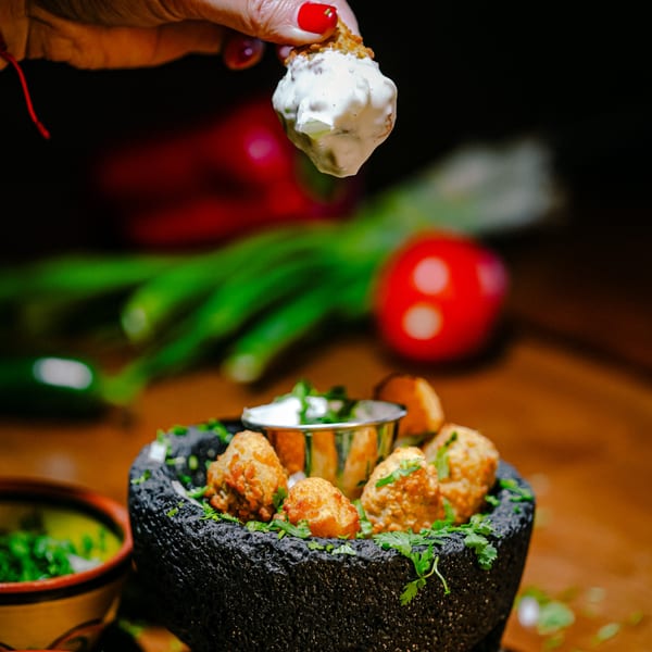 A hand dips a fried Mexican appetizer into a creamy white sauce, served in a dark stone bowl.