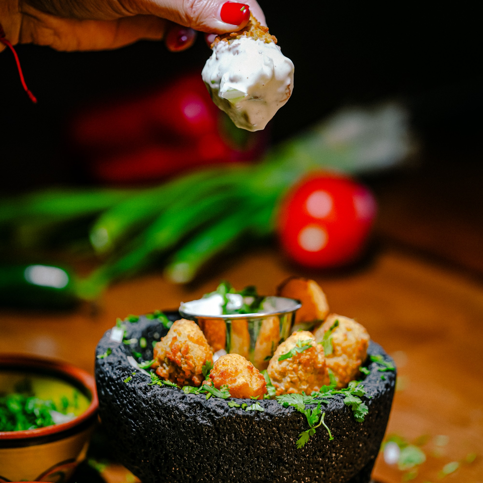 A hand dips a fried Mexican appetizer into a creamy white sauce, served in a dark stone bowl.