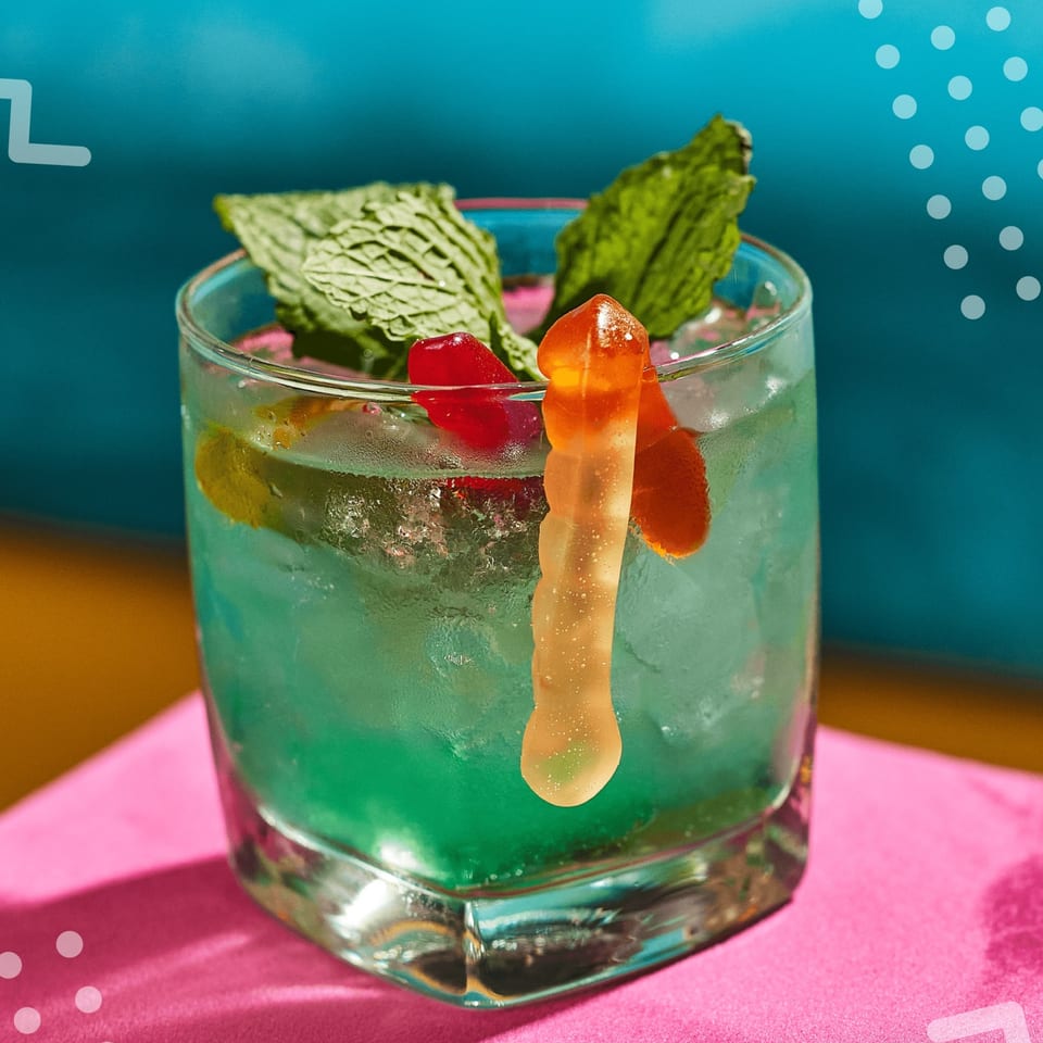 A vibrant blue cocktail garnished with mint, cherries, and a gummy worm on a pink napkin.