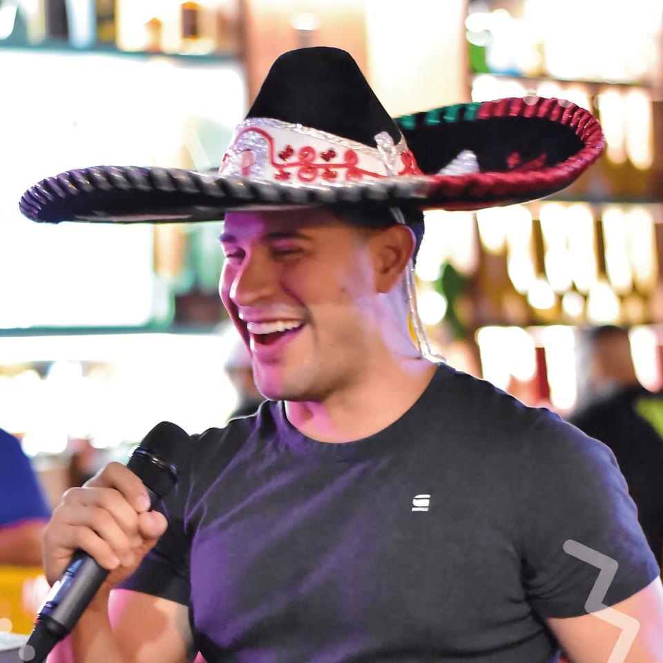 Man in a sombrero and black t-shirt singing into a microphone, laughing joyfully.