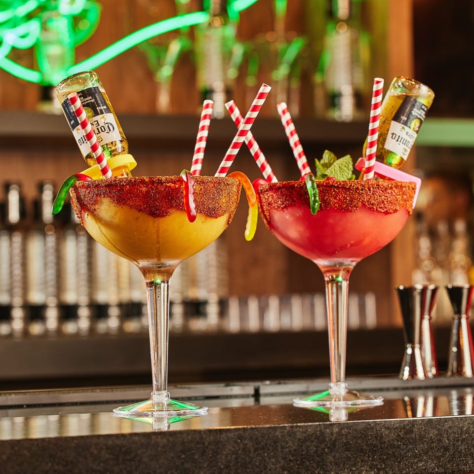 Two colorful Whiskylucan margaritas with beer bottles, candy worms, and chili-lime rims on a bar.