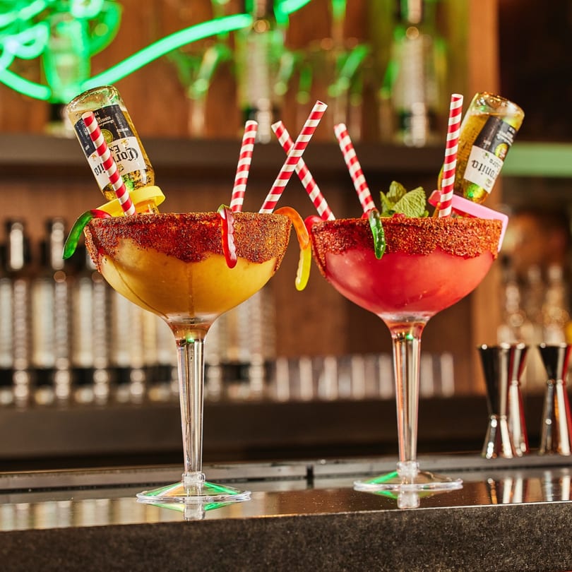 Two colorful Whiskylucan margaritas with beer bottles, candy worms, and chili-lime rims on a bar.