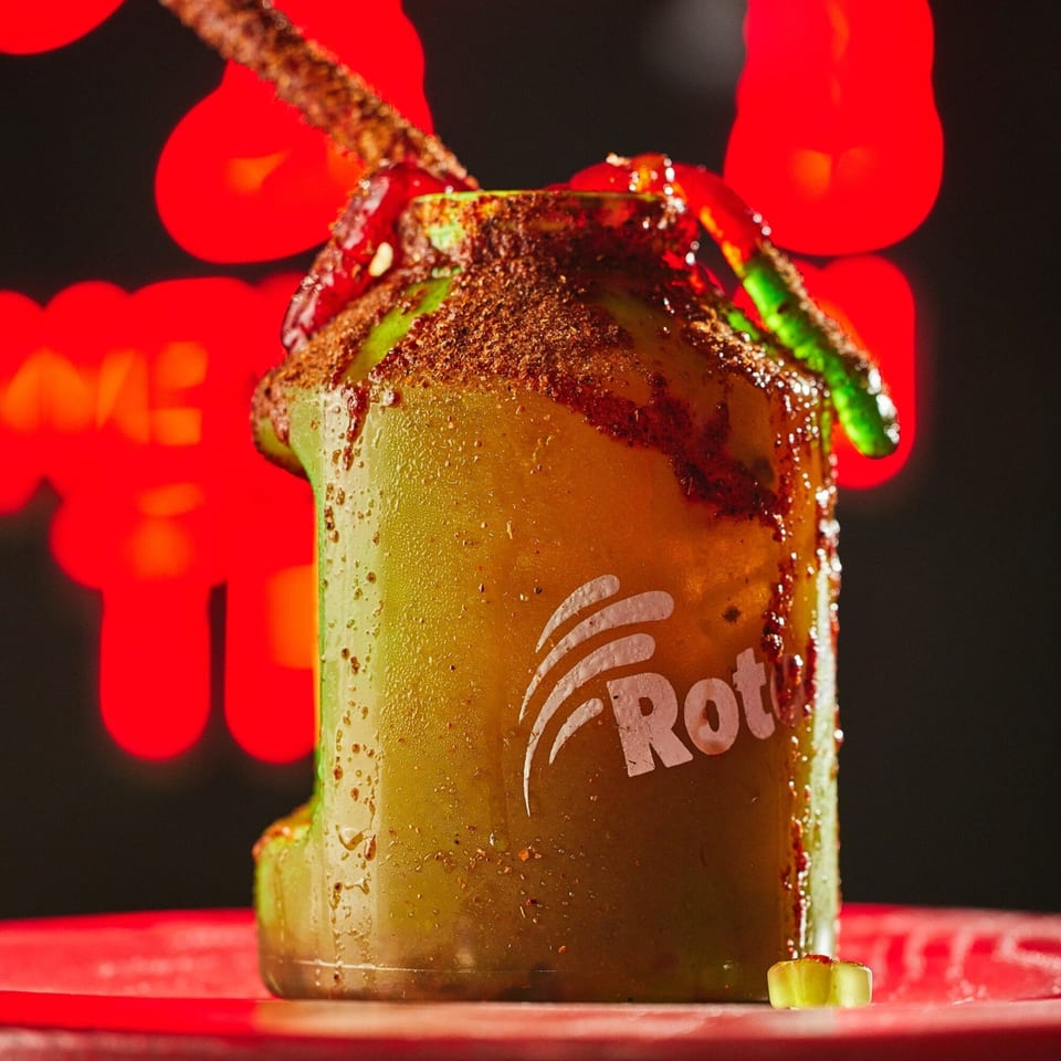 A vibrant green drink in a branded cup, garnished with chili powder, red sauce, and gummy worms.