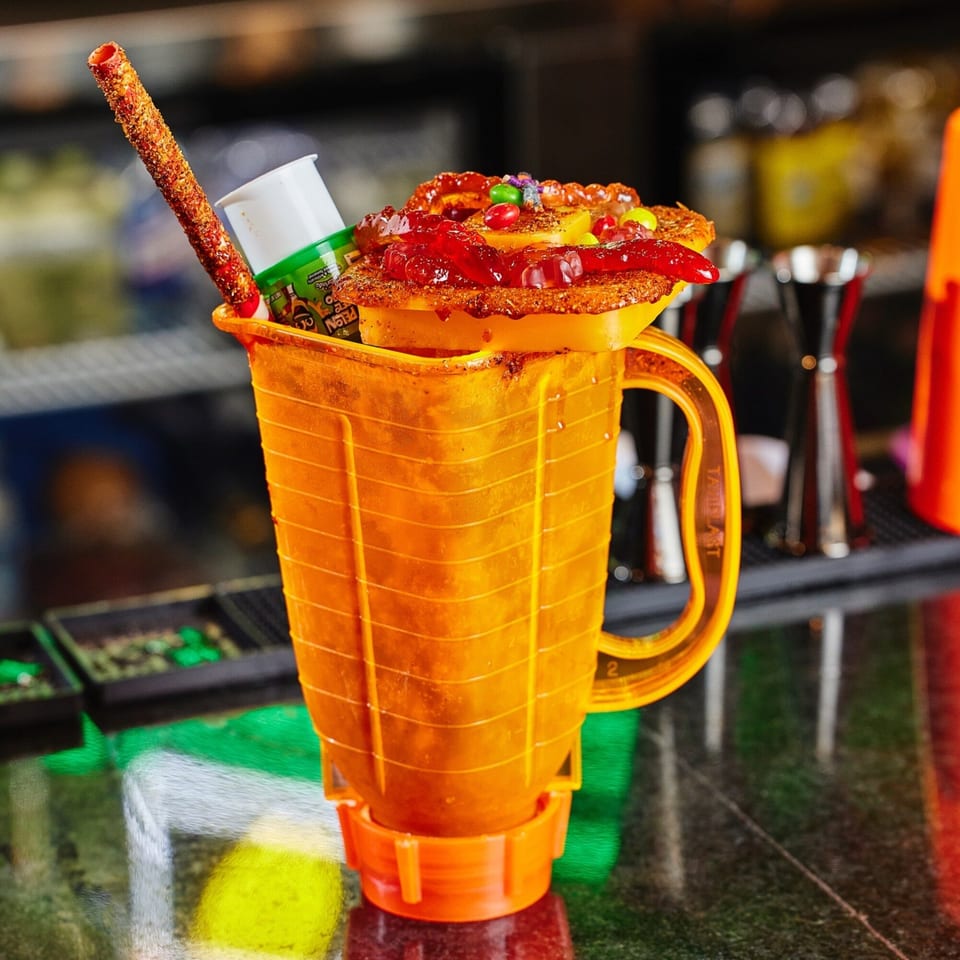 A vibrant orange Mexican drink with candy, fruit, and a chili-dusted straw on a bar.