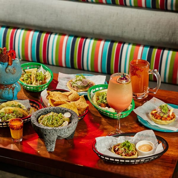 A table laden with various Mexican dishes, including tacos, guacamole, chips, and drinks.