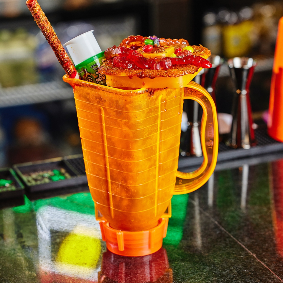 A vibrant orange Mexican michelada, topped with candies, gummies, and a chili-dusted straw.
