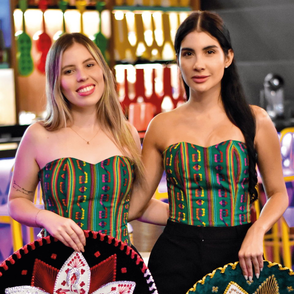 Two women in colorful Mexican-inspired tops hold sombreros in a vibrant restaurant setting.