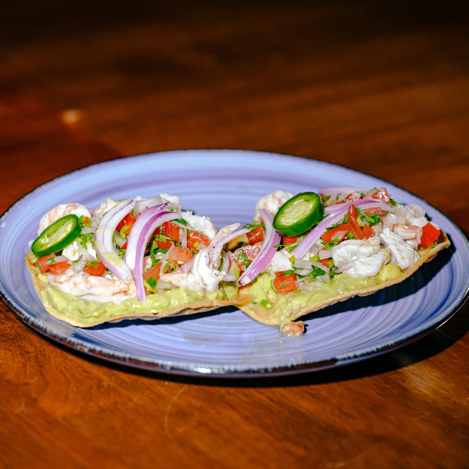 Two Mexican tostadas topped with ceviche, guacamole, red onion, tomato, and jalapeño slices.