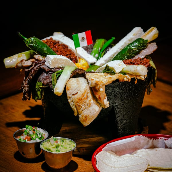 A large Mexican molcajete dish filled with grilled meats, cheese, peppers, and onions, with a Mexican flag.