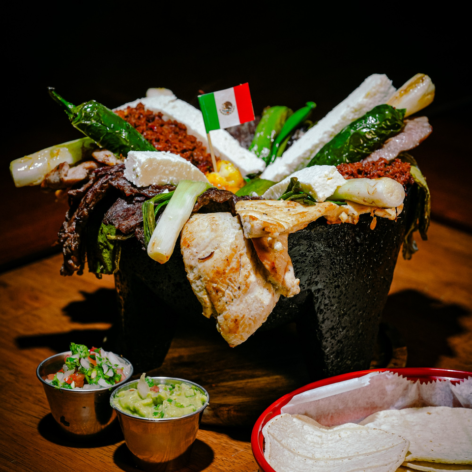 A large Mexican molcajete dish filled with grilled meats, cheese, peppers, and onions, with a Mexican flag.