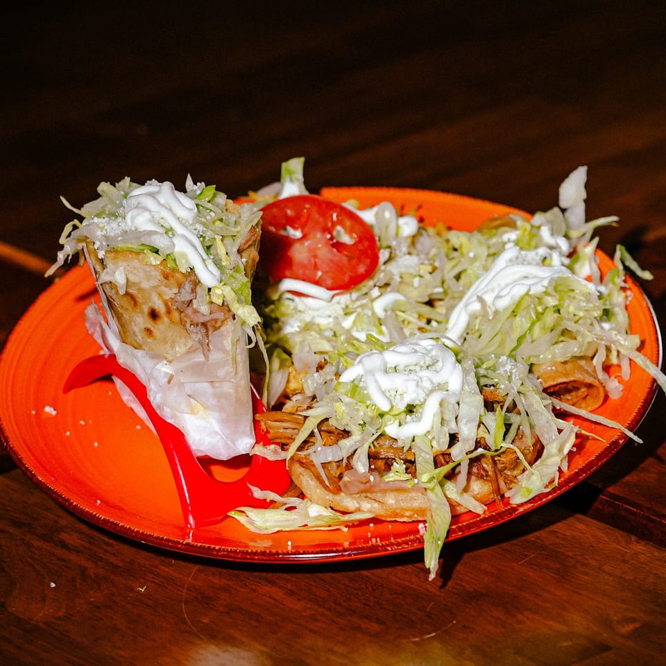 Mexican dish with a burrito, flautas, shredded lettuce, sour cream, and a tomato slice on an orange plate.