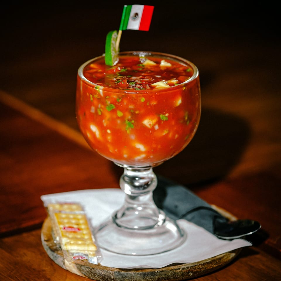 Mexican ceviche in a goblet with a small Mexican flag, lime, and crackers on a wooden board.