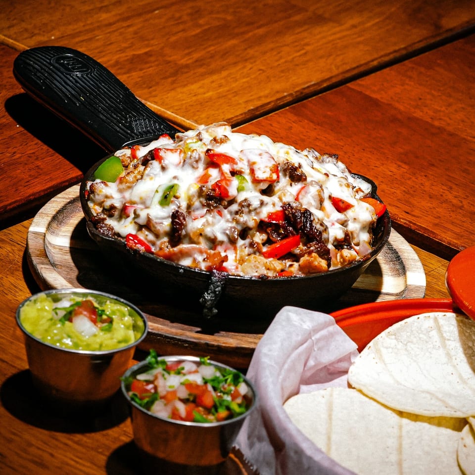 Sizzling Mexican dish with melted cheese, meat, and peppers in a cast-iron skillet, with guacamole and tortillas.